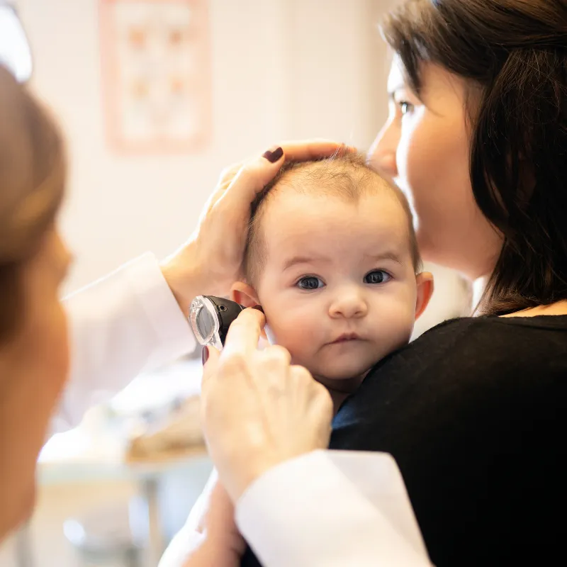 Clinician gently examining a baby’s ear with an otoscope while a parent holds the child